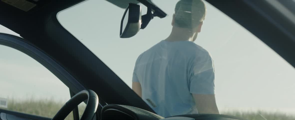 A man stands outside and leans against a car on a sunny day