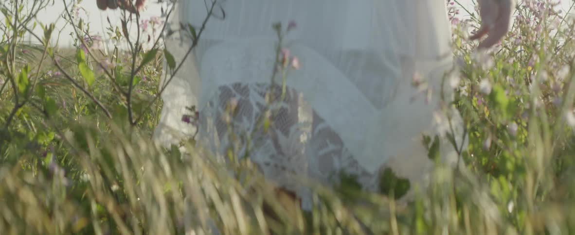 A woman lifts up her white skirt as she walks through tall grass