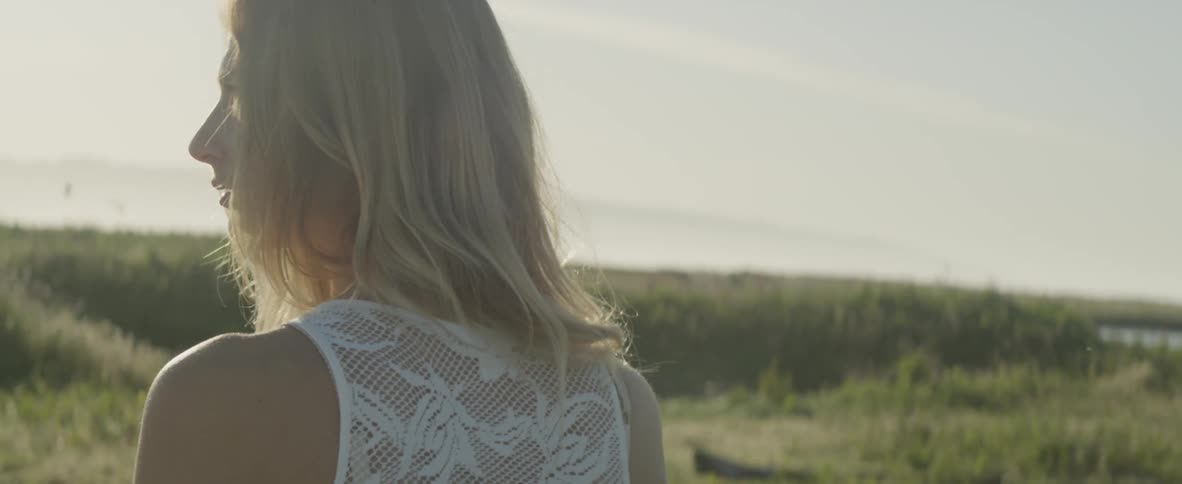A woman walks through a grassy area, turning and looking around on a bright day