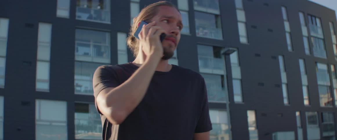 A stressed young man looks worried while talking on his phone outside apartments.