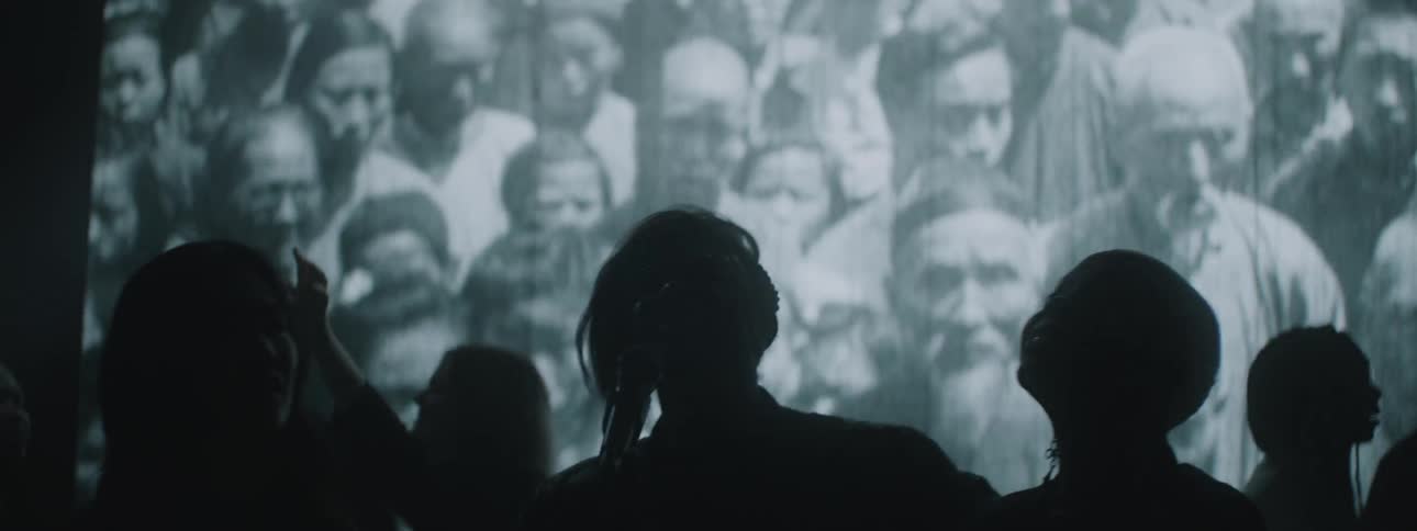 A group of people stand around a microphone singing with a movie screen playing several scenes behind them