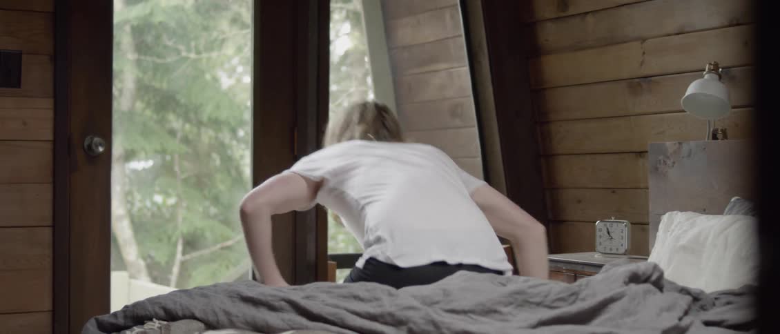 Man waking up and sitting up on the edge of his bed as he looks out at the wilderness outside his window
