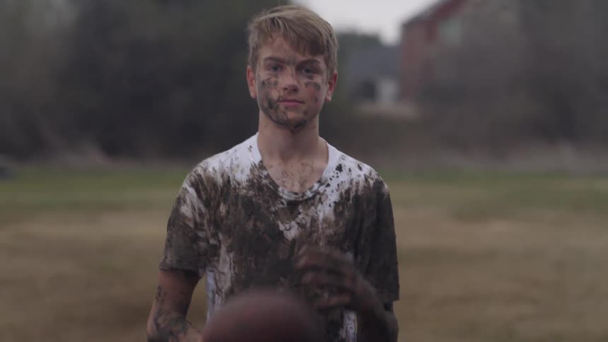 A boy covered in mud tosses a football from one hand to the other