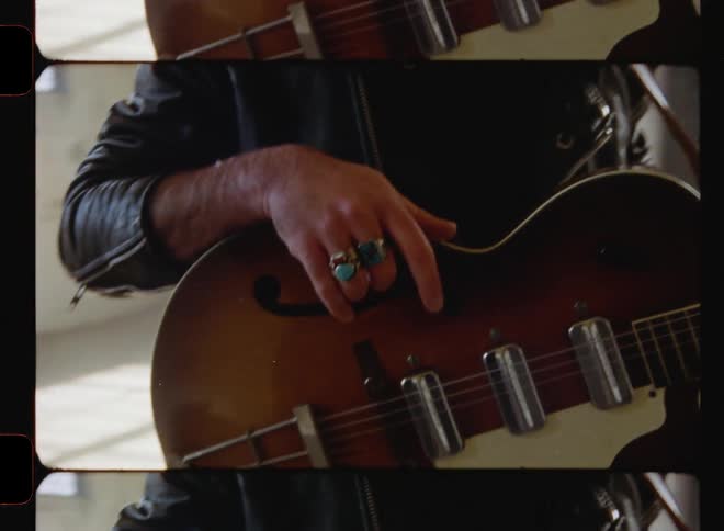 Bearded man wearing a hat stands in an empty room holding a guitar