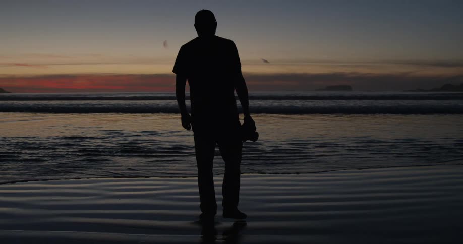 A man holding a camera walks towards the shore at dawn