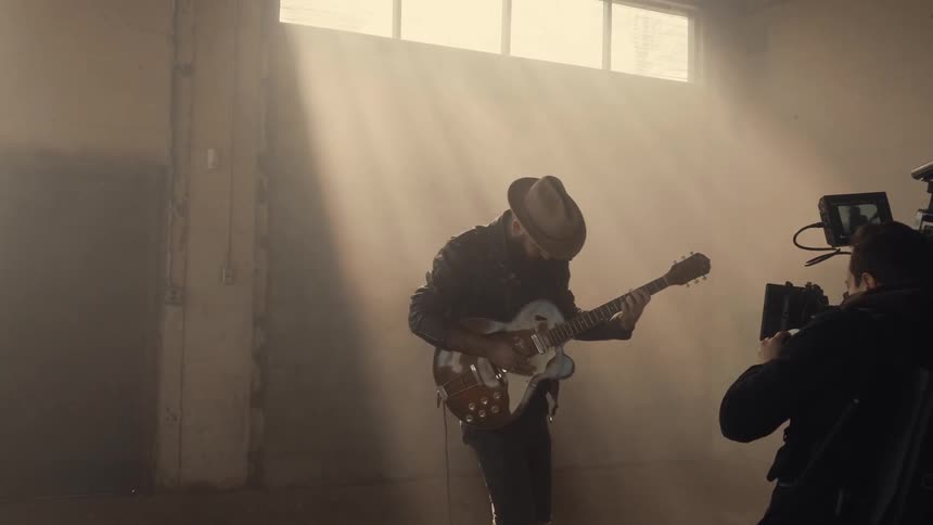 A guitarist is video recorded in a dusty warehouse