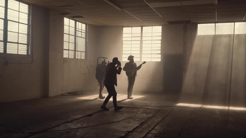 Man playing guitar in a large empty room of a building as two others move around him and filming