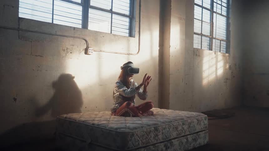 Woman sitting on bed in empty room is wearing virtual reality headset