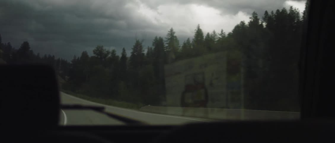 Person driving down a two-lane paved road as seen through the rear windshield on a cloudy day