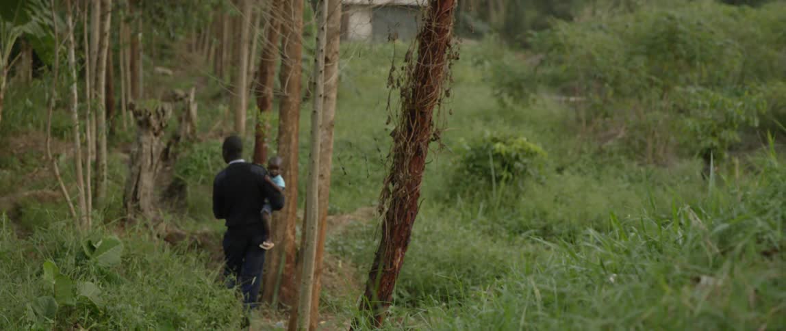 A man carries a small child through a wooded area toward a house