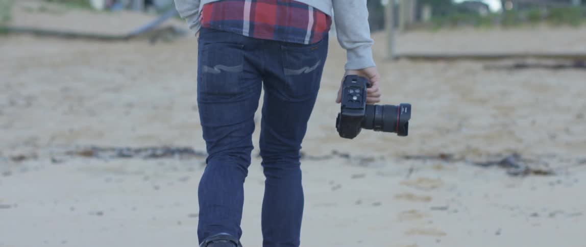 A man carrying a camera walking down a beach towards some buildings