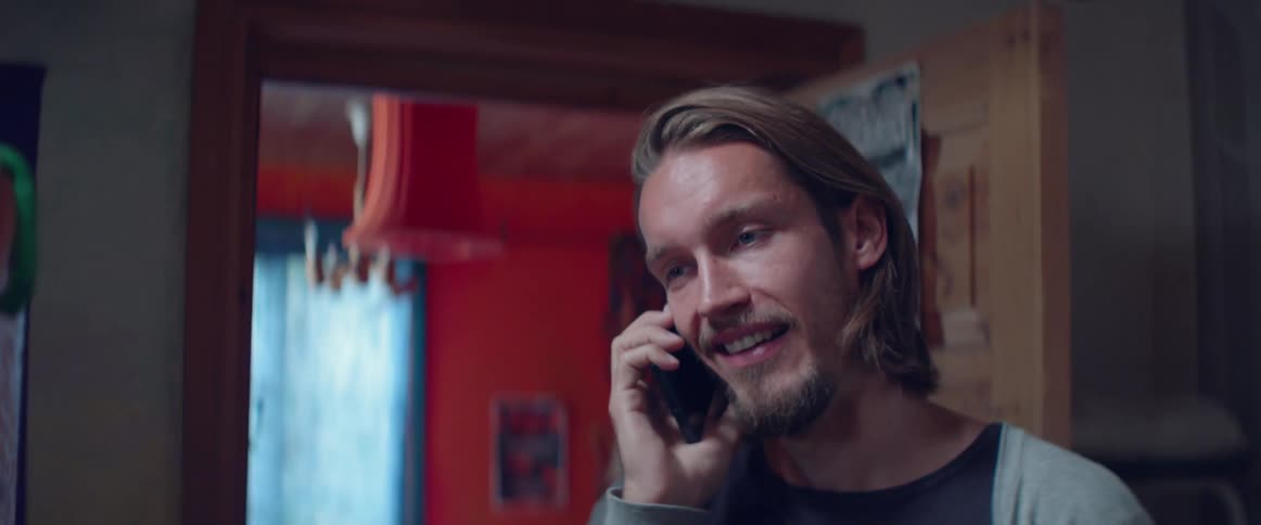A happy young guy laughs during a friendly mobile phone conversation indoors.