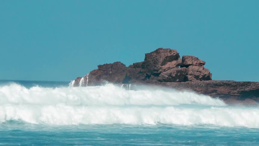 Waves are crashing over a natural rock formation on a sunny day
