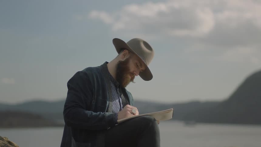 A man is sitting near a lake on a breezy day sketching in a notebook
