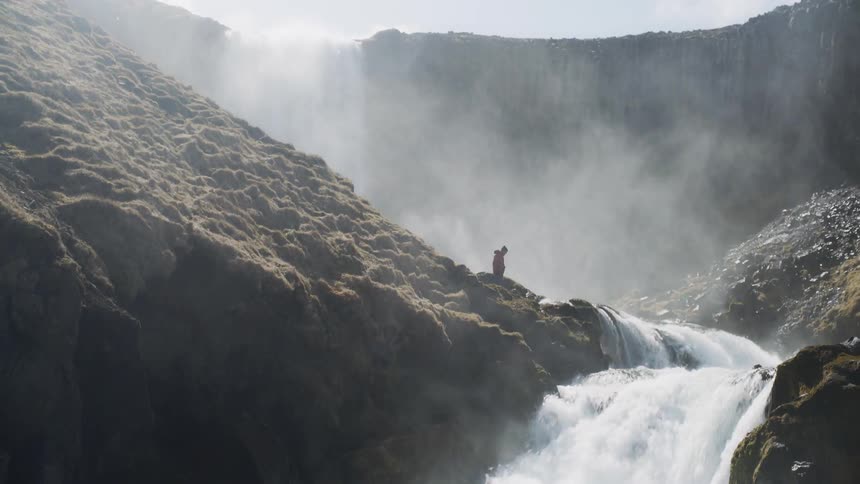 Person standing next to raging waterfall puts their hands in the air