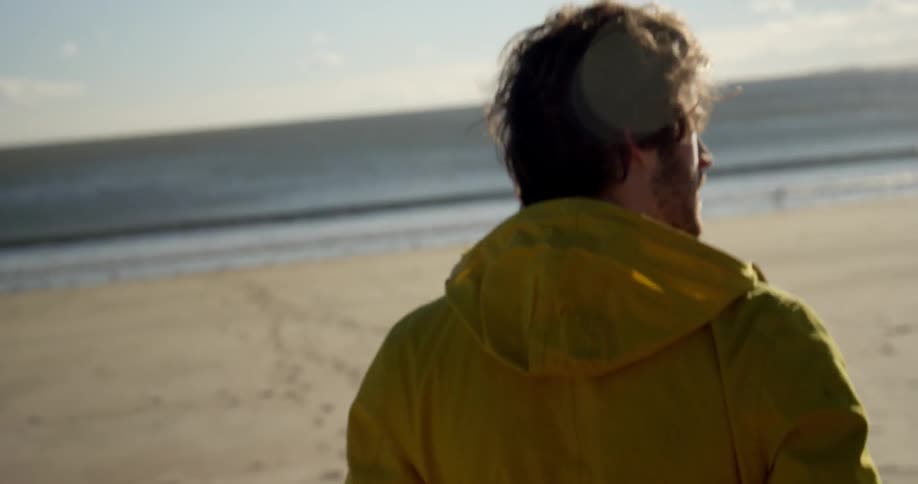 A young, bearded brunette man wearing a yellow jacket walking towards the ocean