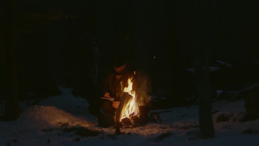 A man sits by a fire in the snow and writes in a notebook