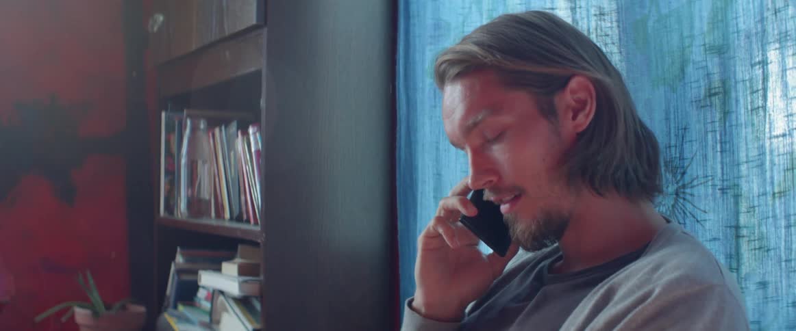 A young guy looks down thoughtfully while having a mobile phone conversation indoors.