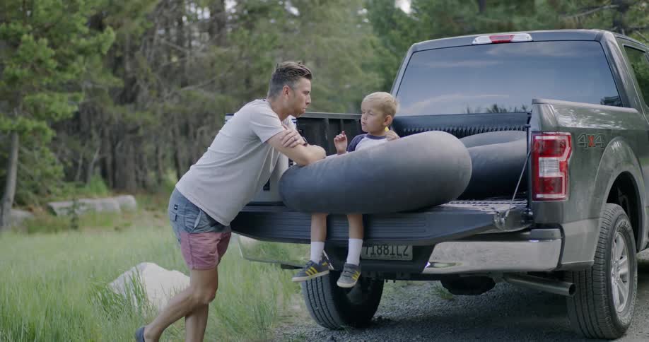 A boy sits in a tube on a truck bed eating while his dad talks to him