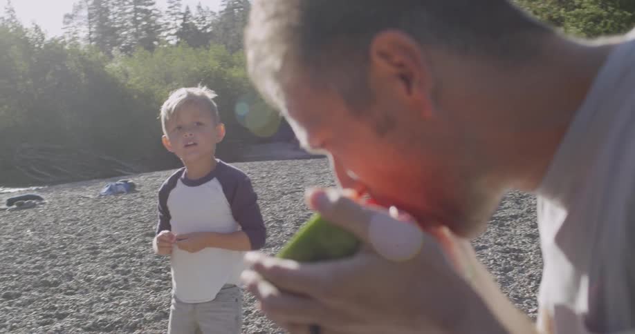 A man eats watermelon while his son throws rocks into the river