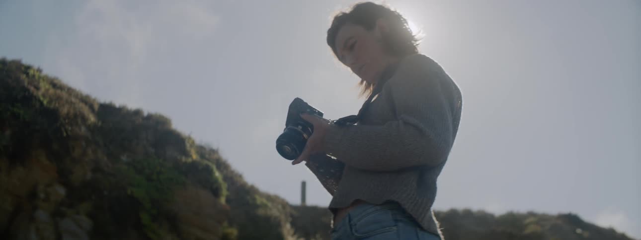 A woman is looking at the back of her camera as she stands outdoors in sunlight