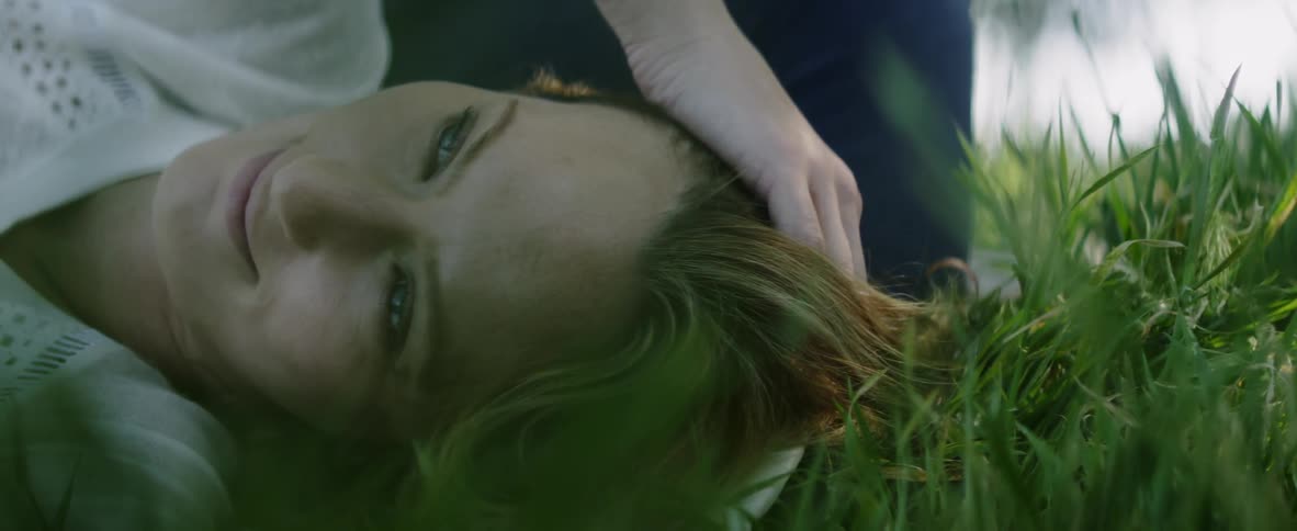A woman laying in the grass in daylight is touching her head and hair
