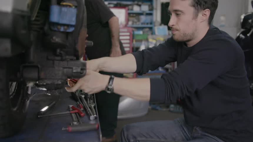 A man is working inside a garage in the daylight repairing a moped