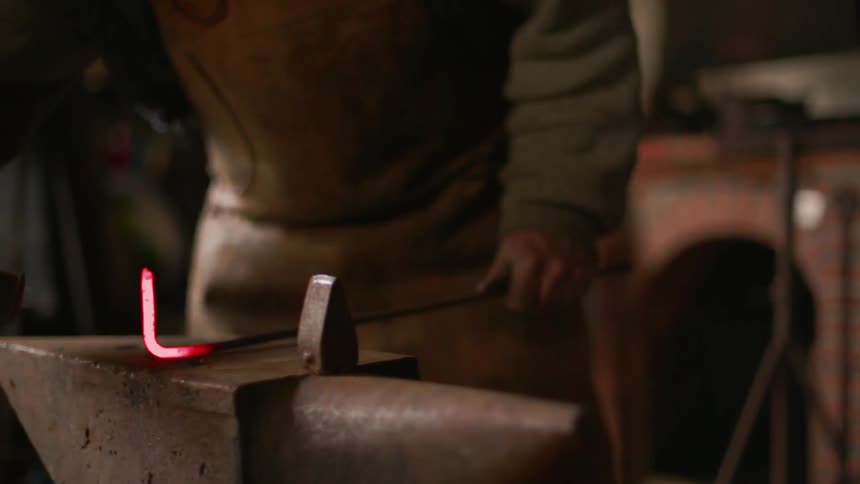 An artisan is forming a metal rod as he works in a shop in daylight