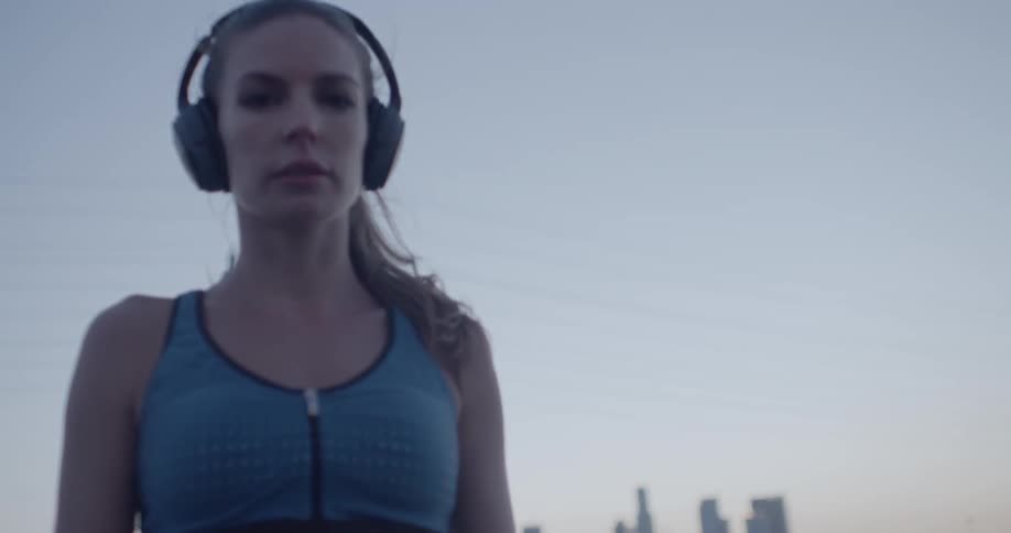 A female athlete is standing outside listening to her headphones at dusk