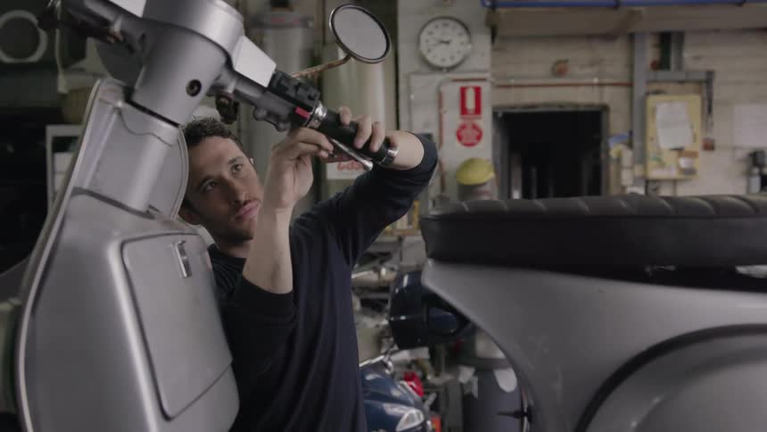 A man is working on a moped inside a garage in daylight