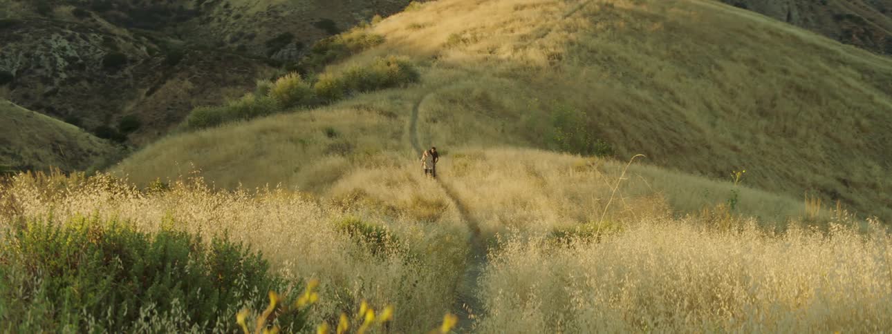 Two people walk on a small path through grassy hilly land