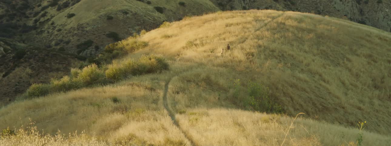 Two people walk on a small path through grassy hilly land