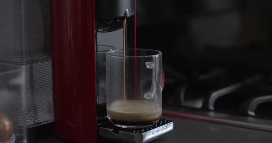 Coffee pours out of a red coffee maker into a glass mug