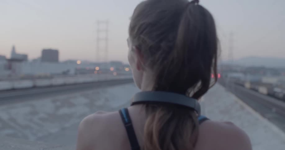 A woman with headphones stands on a city bridge at dusk