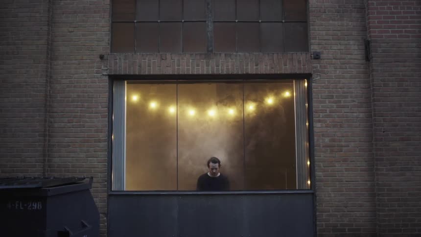 A man stands still in front of a large window as smoke surrounds him