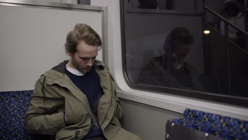 Man with a khaki coat sits on a train and looks down at his feet at night