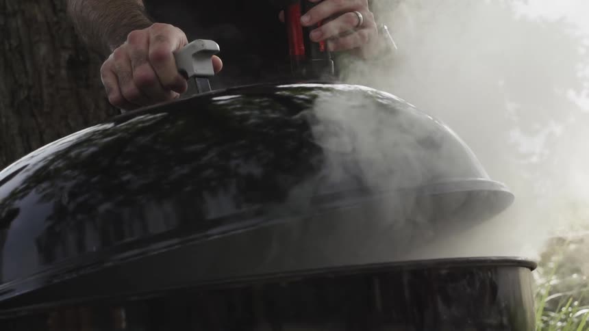 A person lowers the grill lid as smoke rises