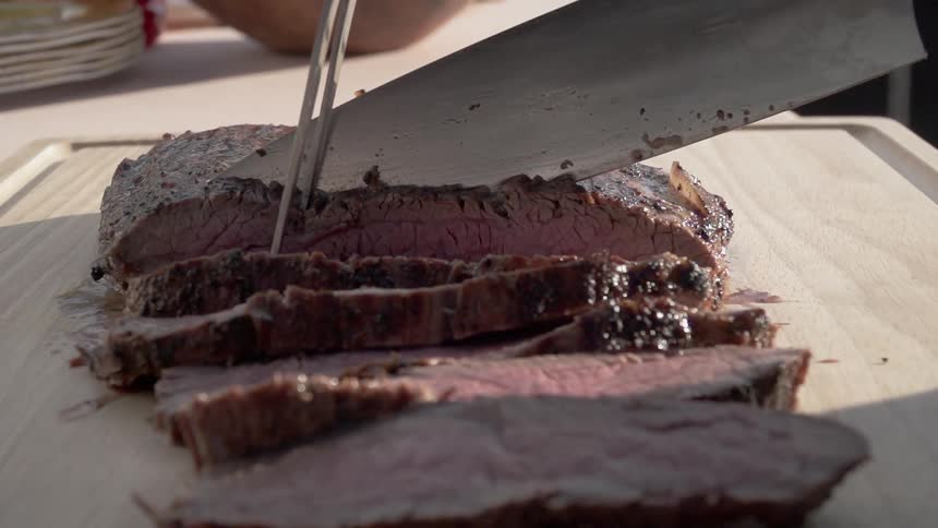 A slab of roasted meat is carefully carved on a cutting board outside