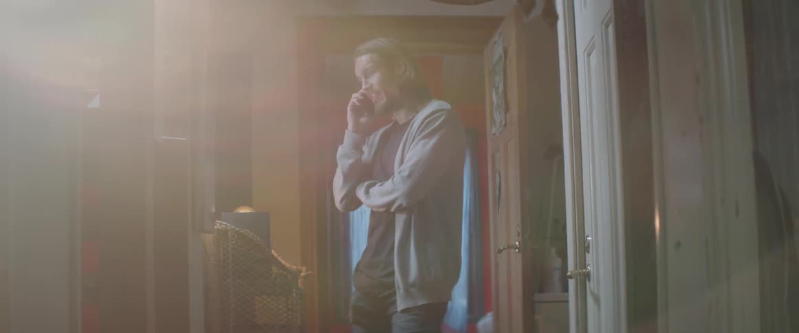 A young guy wearing a grey cardigan stands indoors speaking on a smartphone.