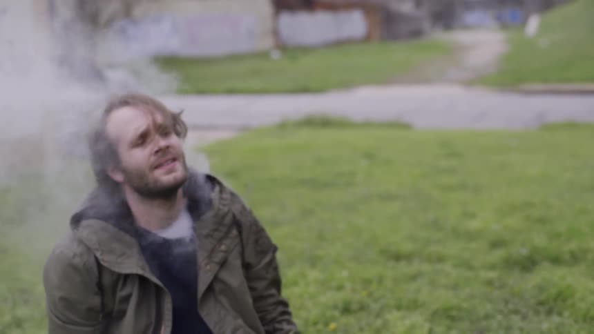 A man falls to the ground sobbing as smoke rises from within his coat and surrounds his head