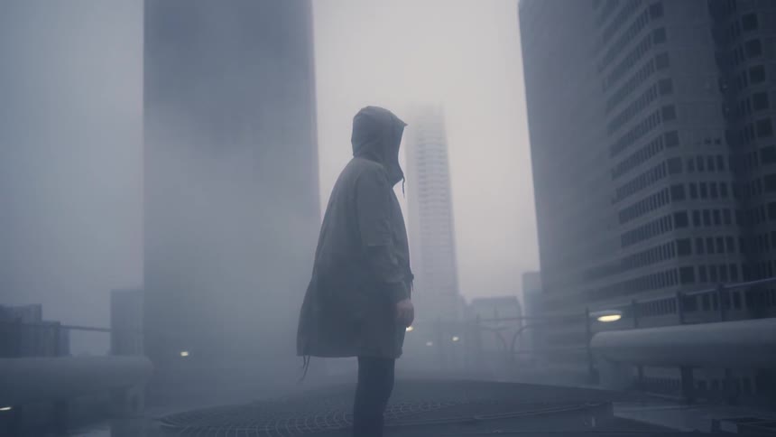 A man wearing a stadium-length coat standing outside in an urban area as fog blows around him