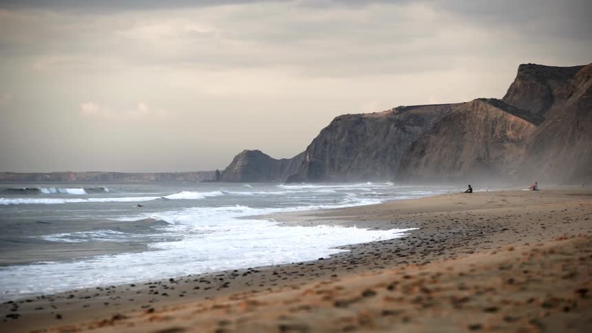 A person sits on the beach while a surfer paddles out into the waves