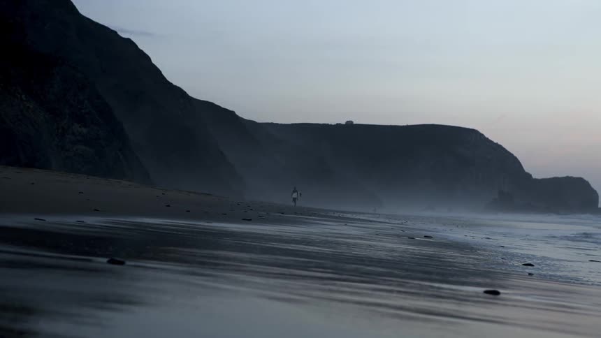A surfer exits the beach as the waves splash on the shore
