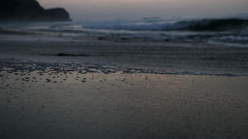 A wave is lapping the sand on a beach at dusk as a surfer heads out in the distance