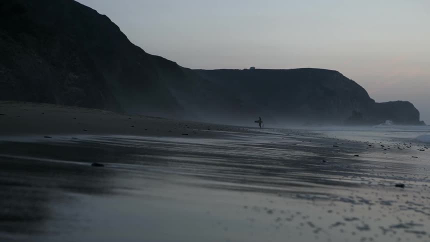 A surfer is setting his board down on the sand at a beach at dusk