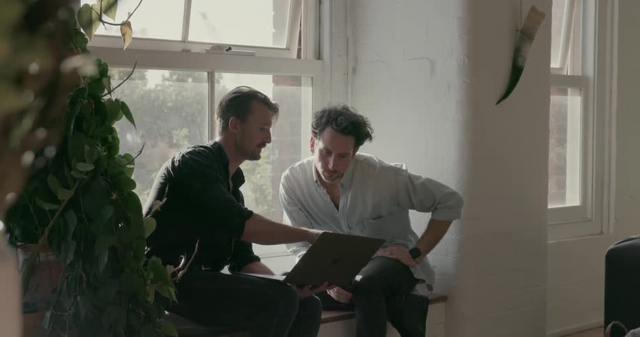 Two men look at a laptop while sitting next to a window
