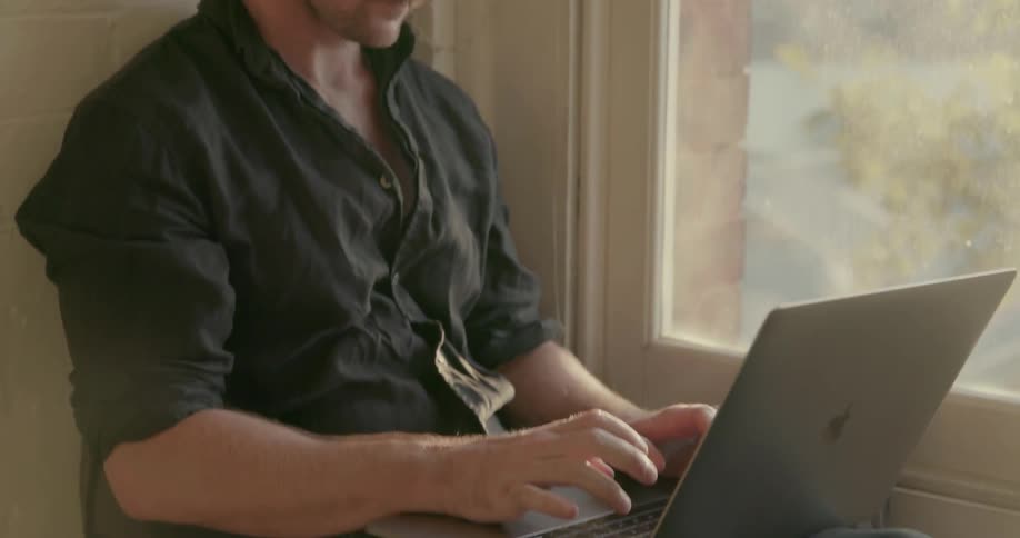 A man types on a laptop while sitting next to a window