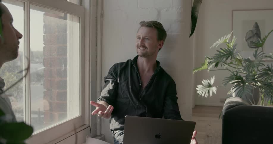 Two men sit on a window sill talking on a sunny day
