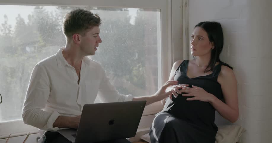 Man types on a laptop and talks to a pregnant woman as they sit