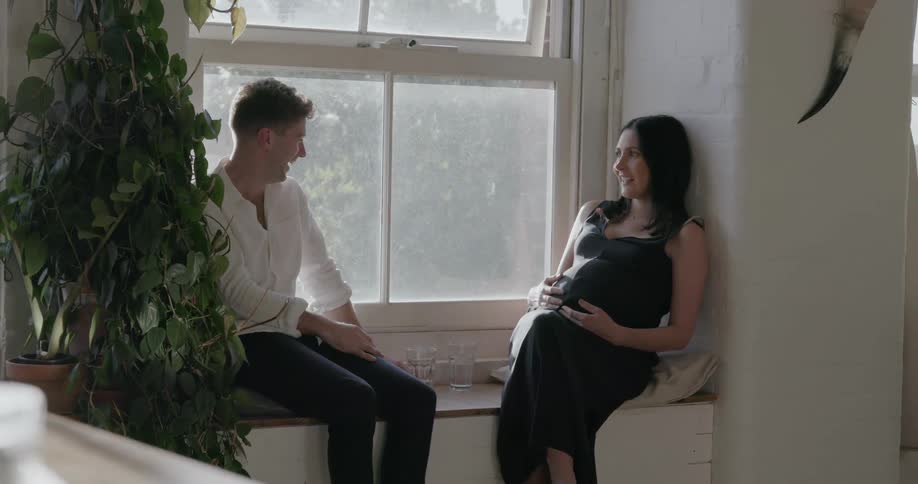 A man and his pregnant wife sit on a window seat talking and laughing